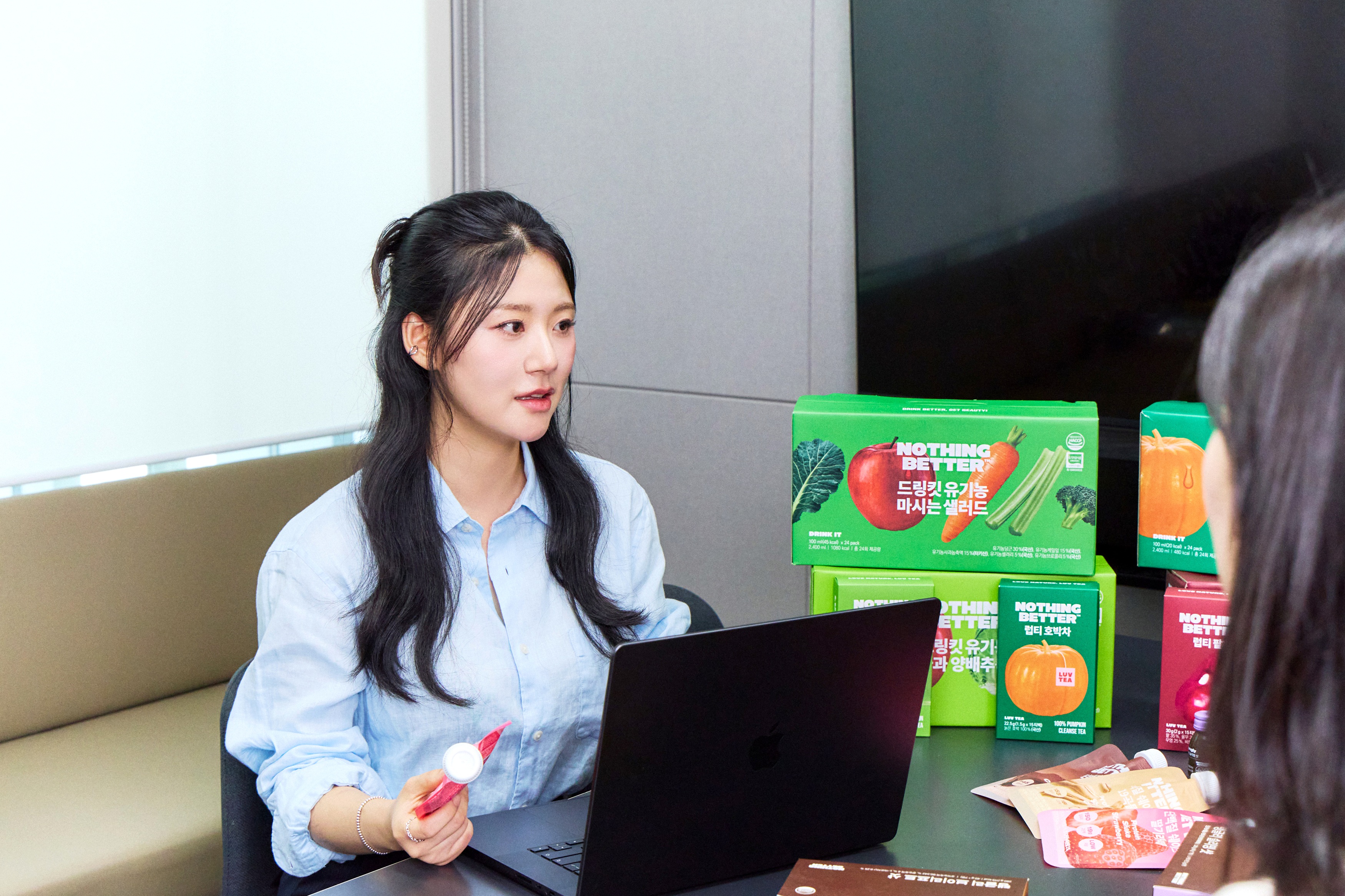 A woman holds a pink drink bottle beside an open laptop and colorful beverage boxes.