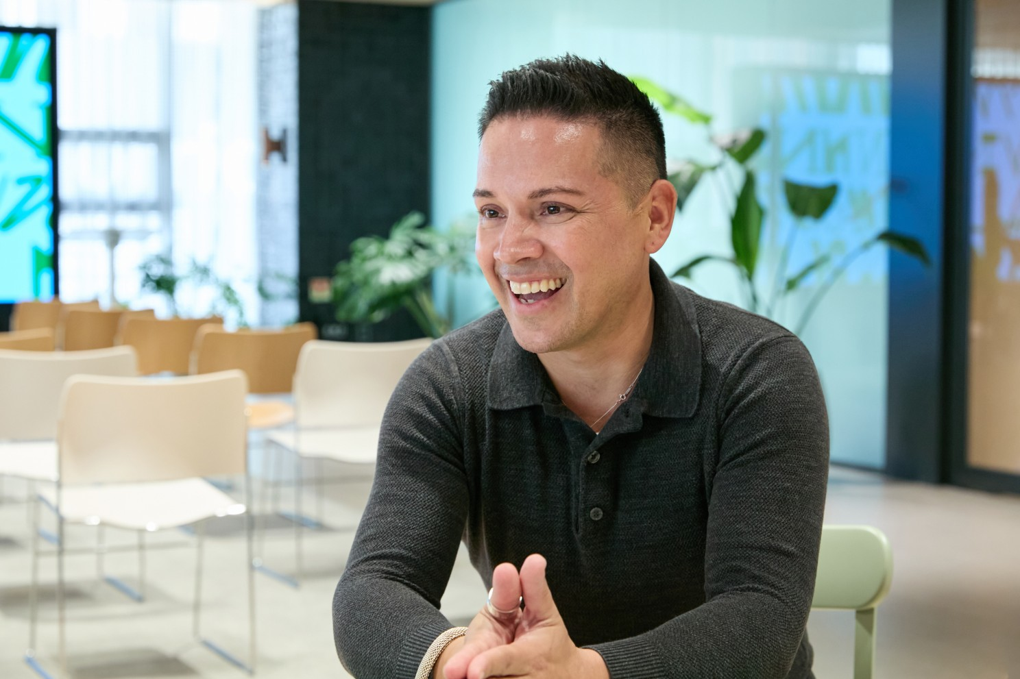 A man laughs while seated in a bright lounge with tables, chairs, and plants.