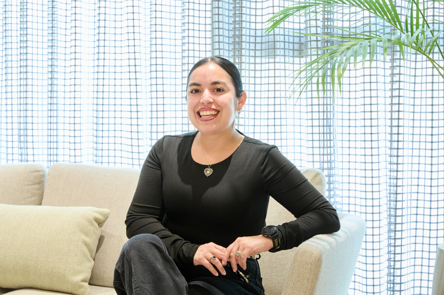 A smiling woman in a black top sits on a couch before a bright grid-patterned window.