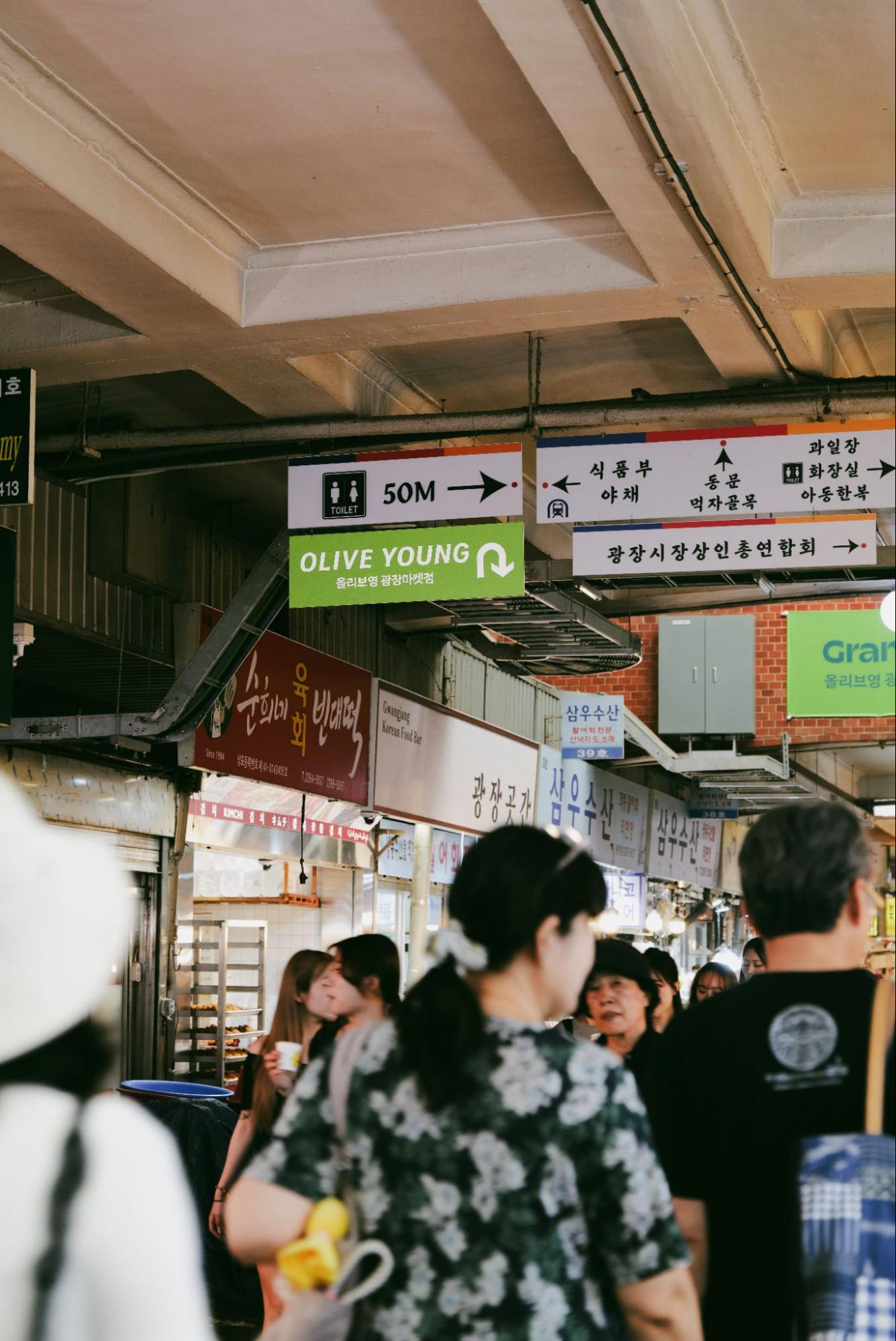 韓国語の案内看板が並ぶ市場の通路を、多くの買い物客が行き交っている。
