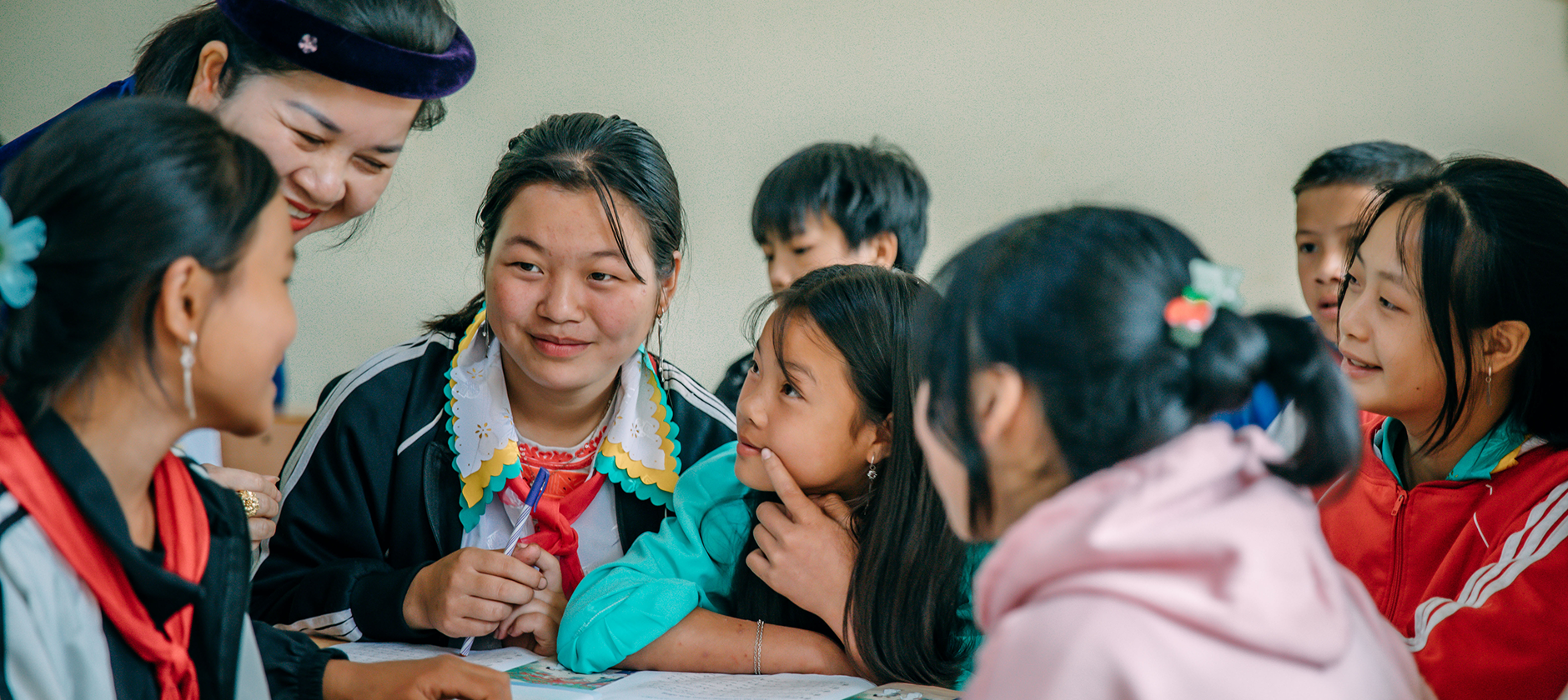 A woman and several children gather around a table, talking and writing.