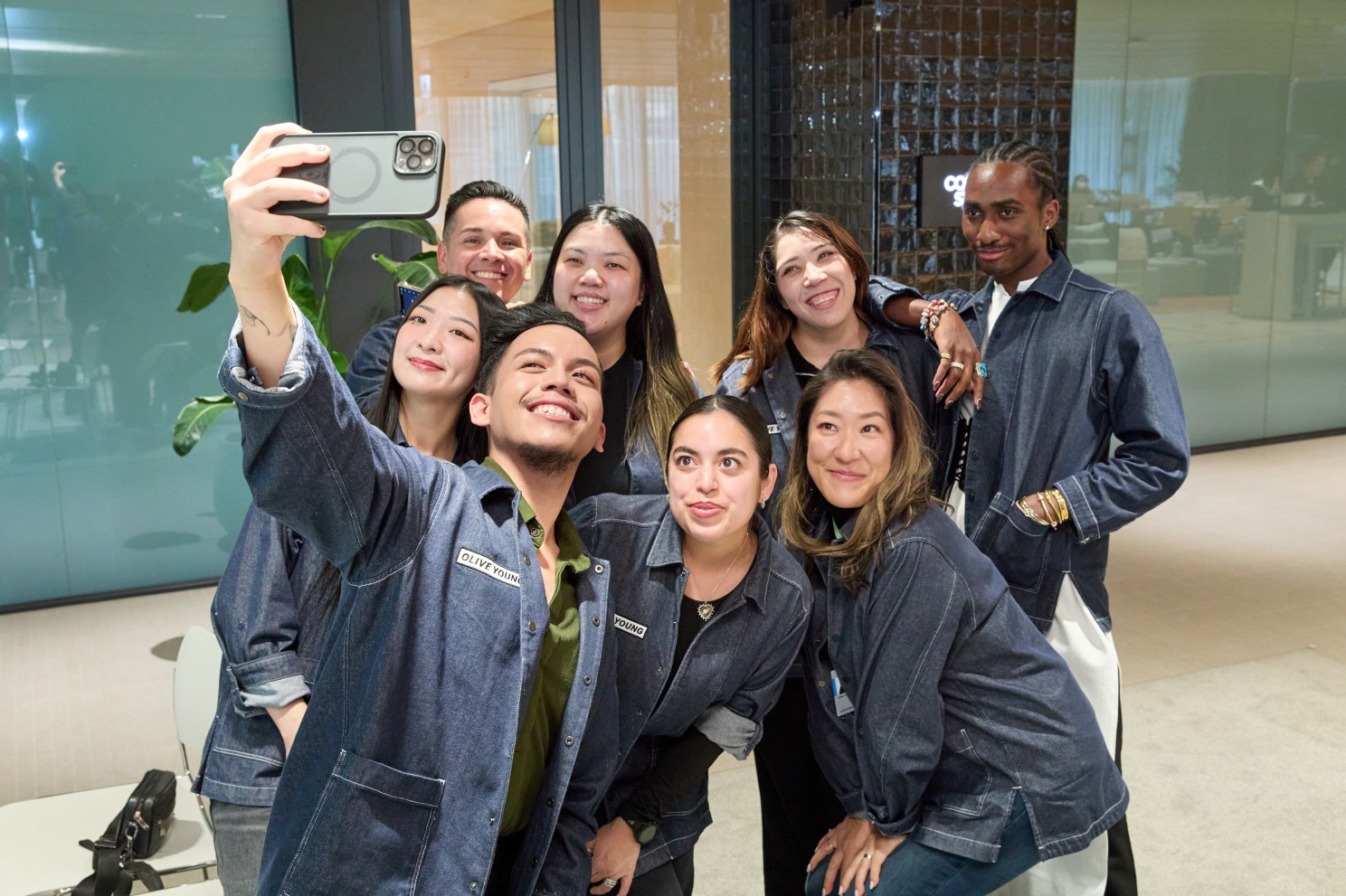 Eight people in denim jackets pose for a selfie in a modern lobby.