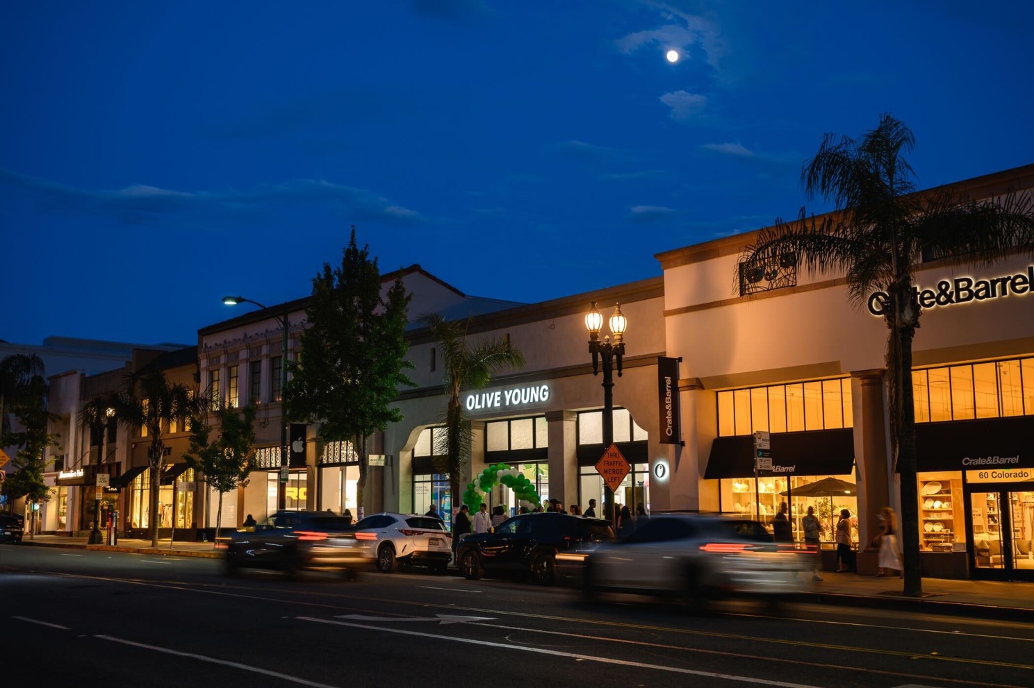 Olive Young and Crate&Barrel storefronts glow along a busy street at dusk as cars pass.