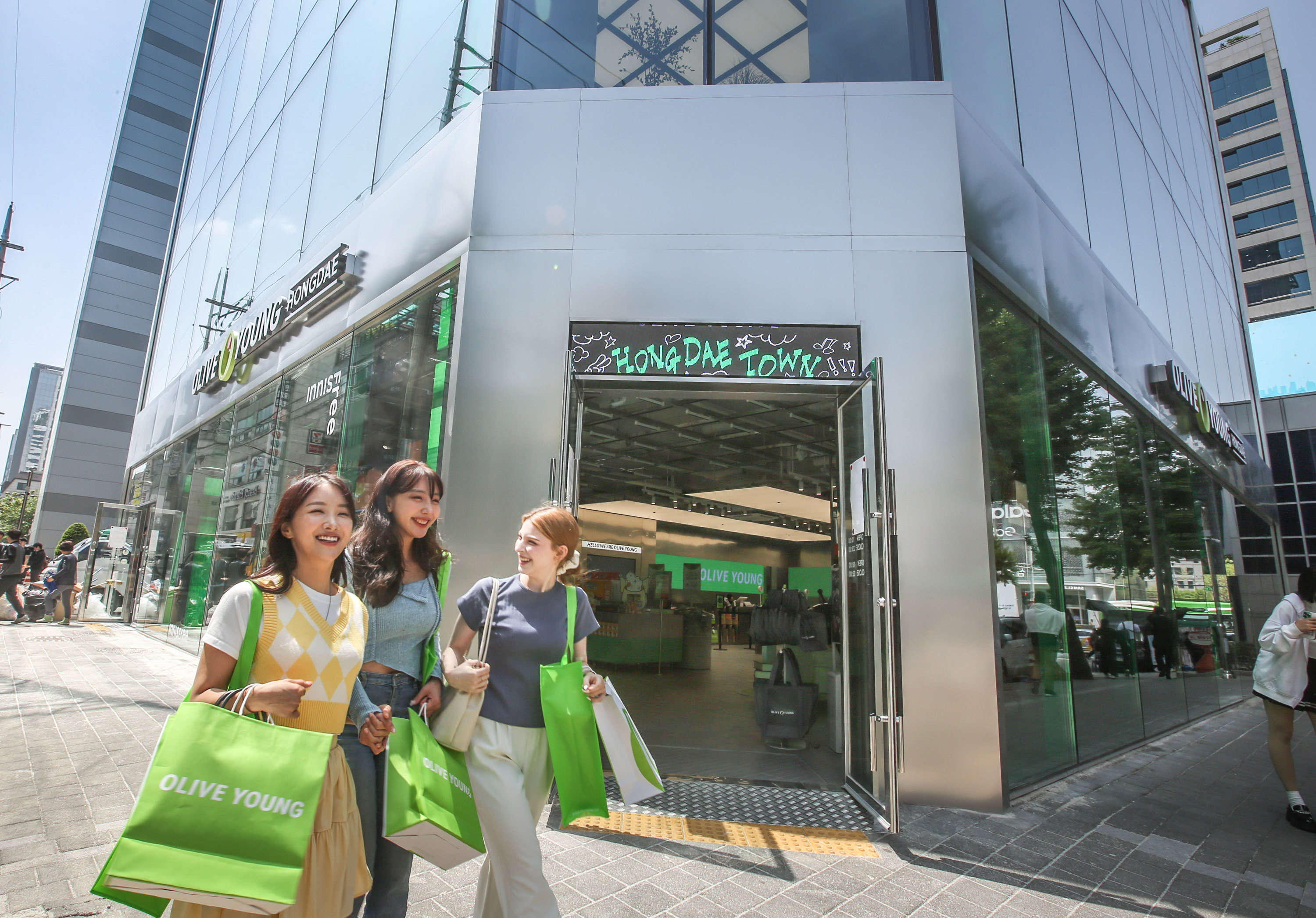Three women smile while holding green shopping bags in front of an Olive Young store.