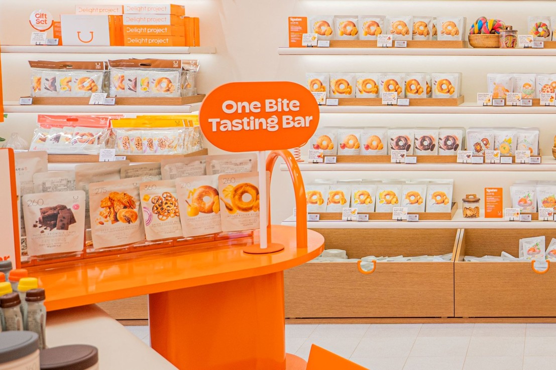 A bakery display features shelves of pastries behind an orange "One Bite Tasting Bar."