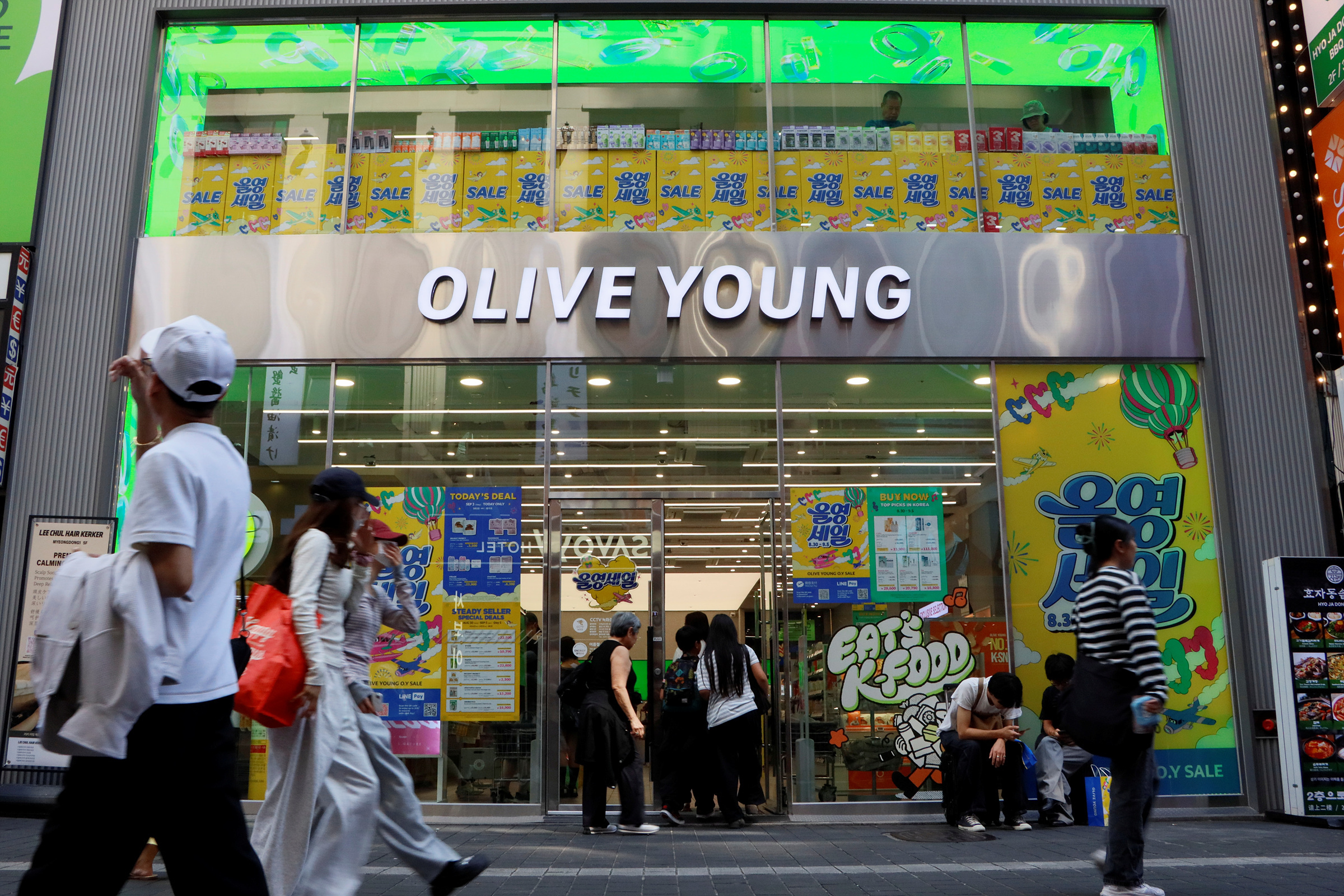Shoppers walk past an Olive Young beauty store covered in colorful sale advertisements.