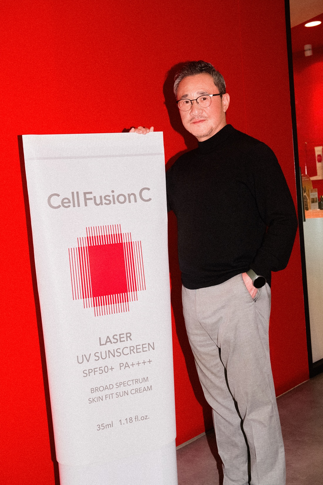 안경을 쓴 남성이 빨간 배경 앞에서 ‘Cell Fusion C’와 자외선 차단제 문구가 적힌 포스터를 들고 있다.