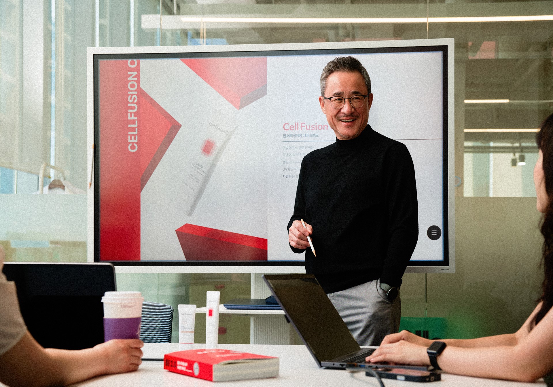 남성이 펜을 들고 셀퓨전 로고가 보이는 대형 화면 앞에 서 있으며, 주변 사람들이 노트북을 사용하고 있다.