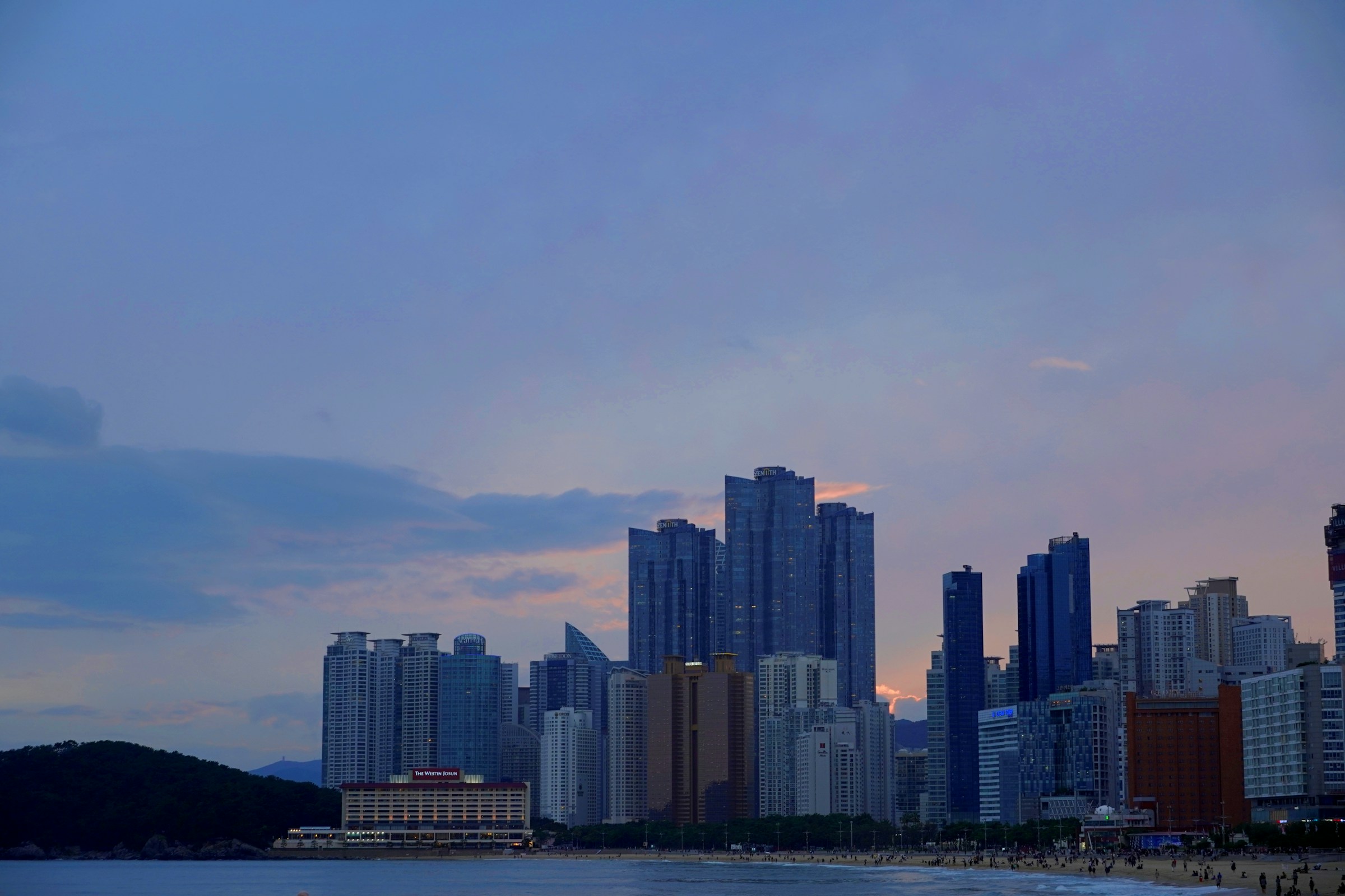 夕暮れの海岸越しに、高層ビルが立ち並ぶ都市の街並みが広がっている。