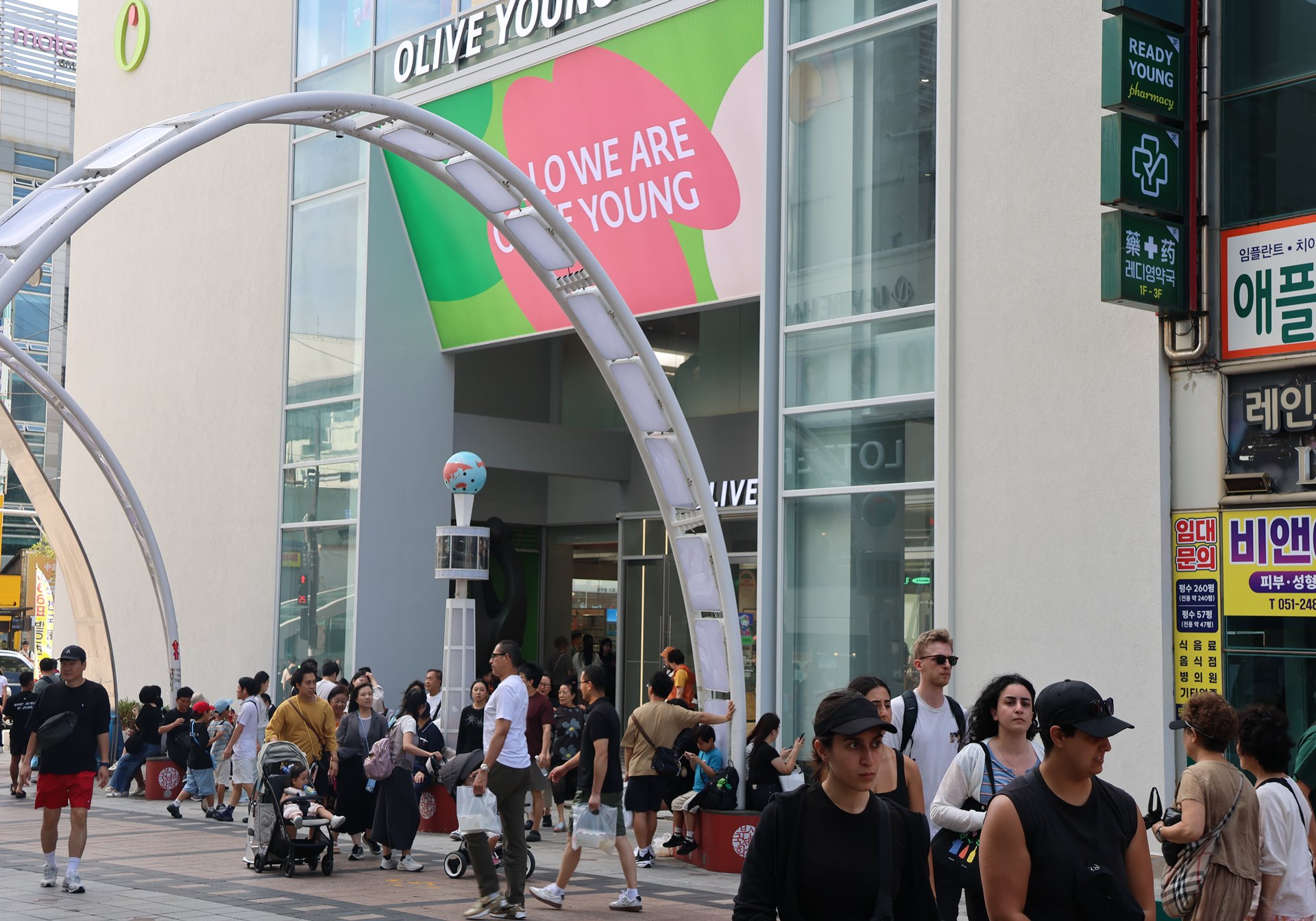 People gather outside an Olive Young store beneath a large curved metal arch.
