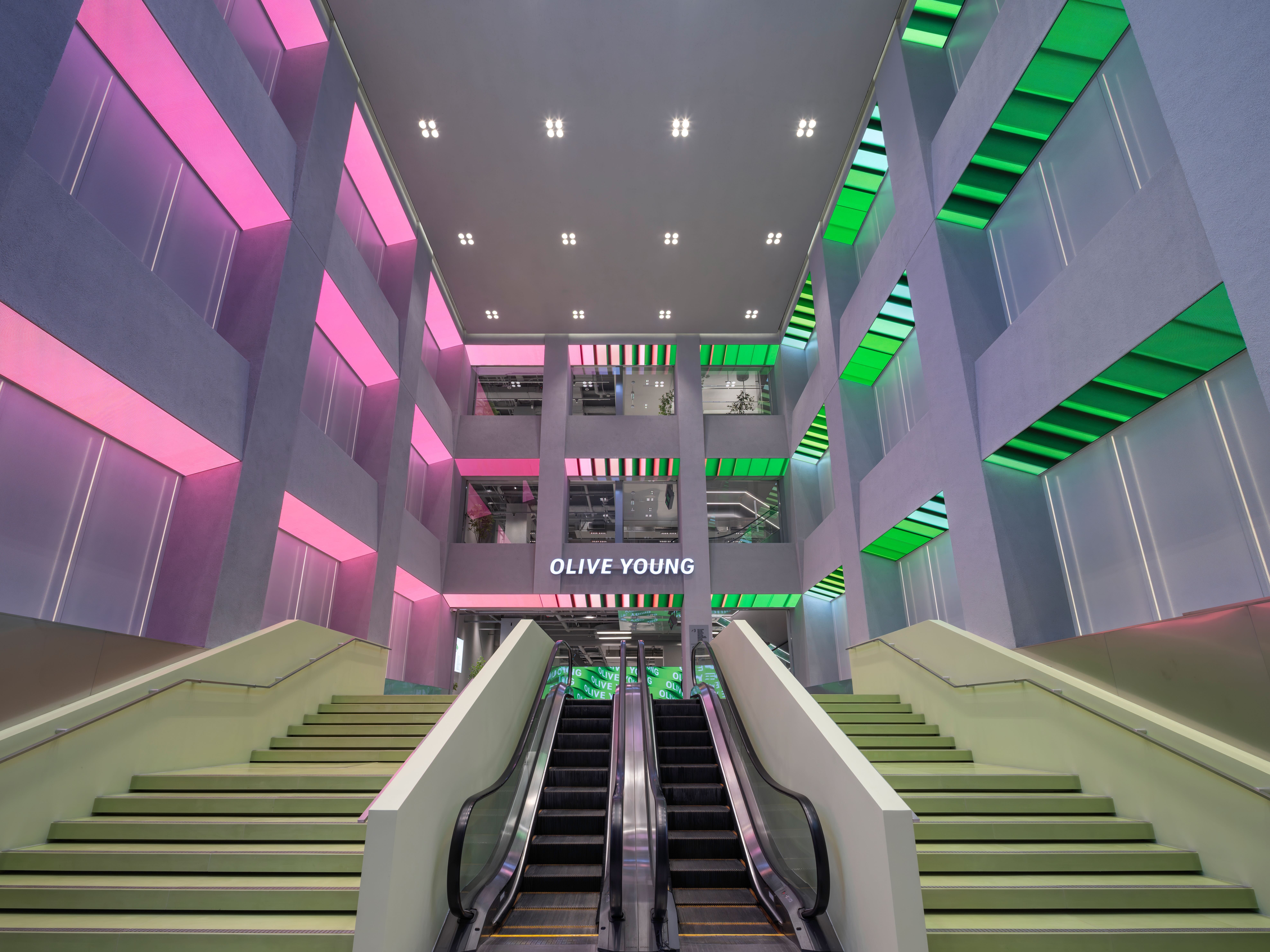 A colorful Olive Young store interior with escalators, stairs and pink and green lighting.
