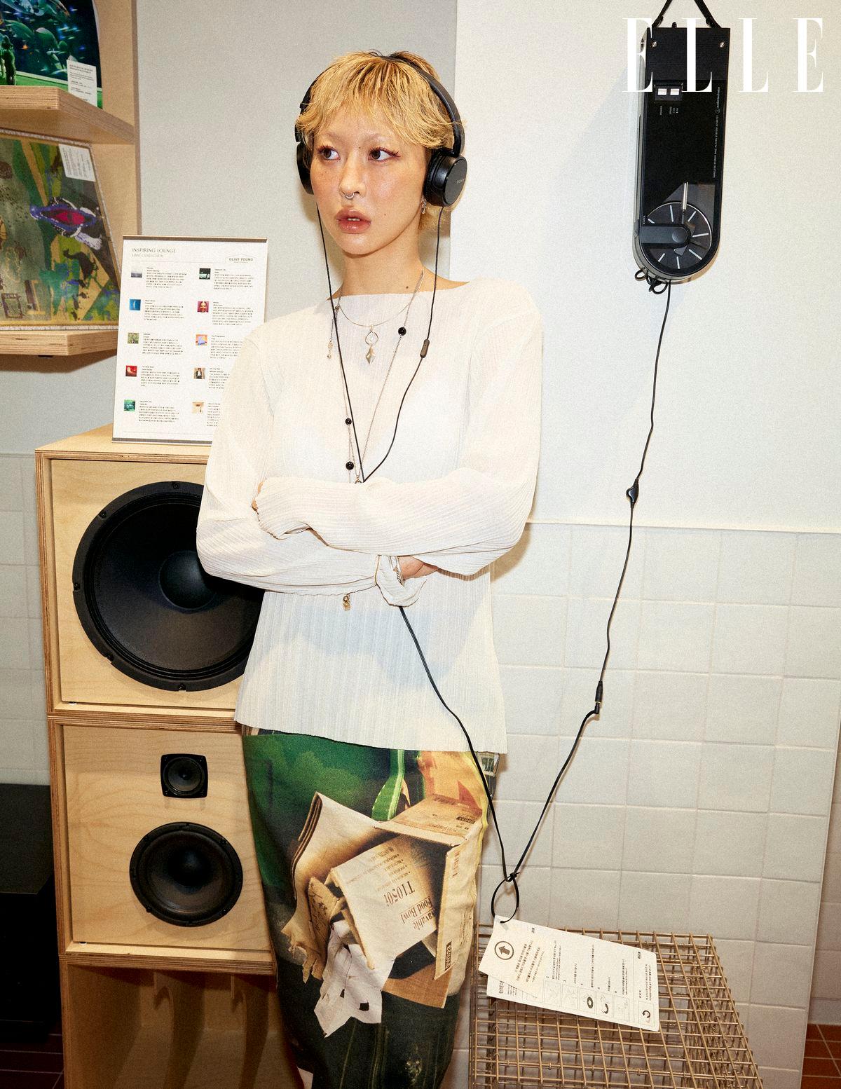 Person wearing headphones stands with folded arms beside large speakers in an exhibit.