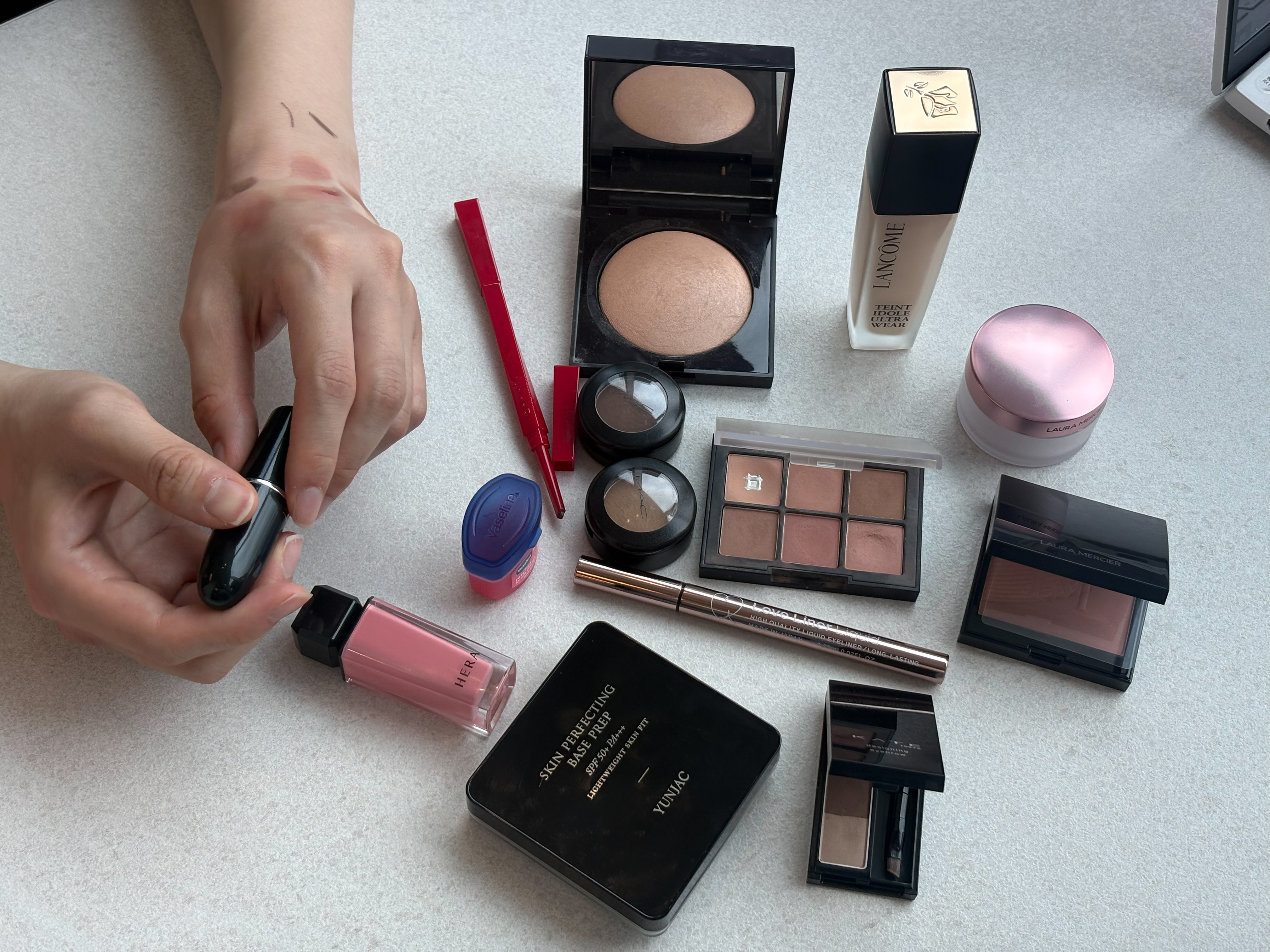 A hand holds a makeup tube beside palettes, lipstick, eyeliner, and other cosmetics.