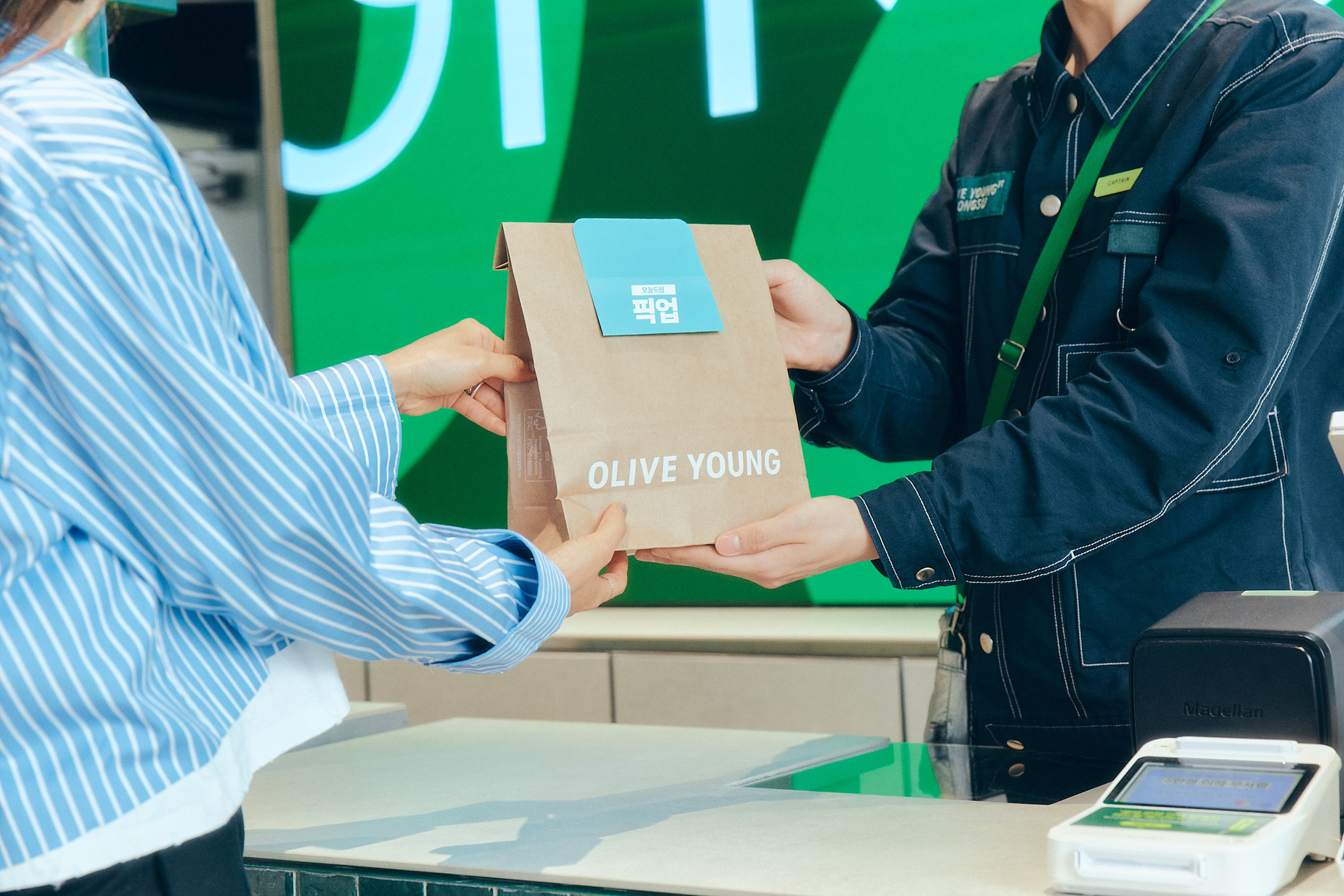 A shopper receives an Olive Young paper bag from a uniformed store worker.