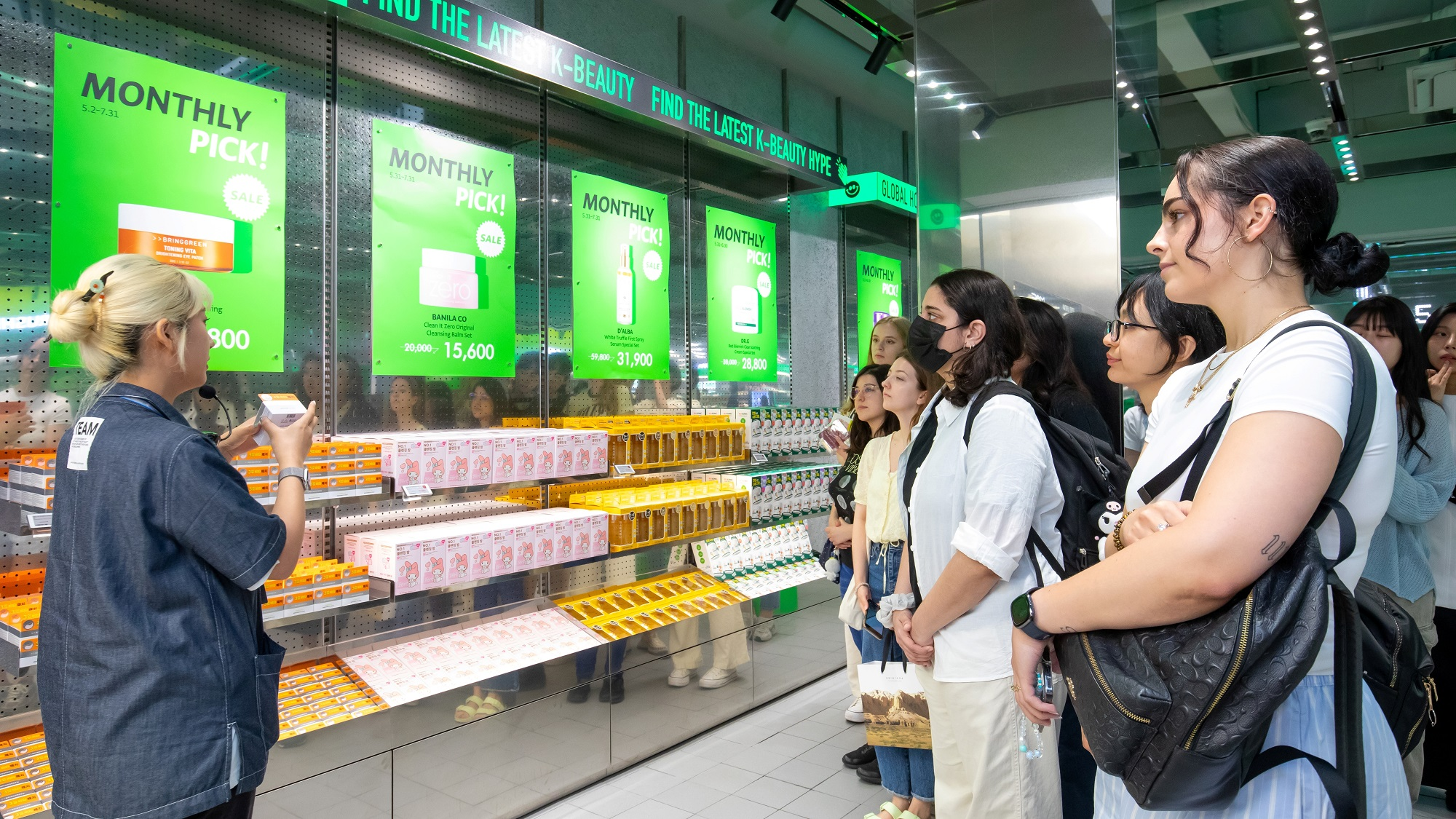 韓国コスメ店で、店員が商品を手に取り、客たちに説明している