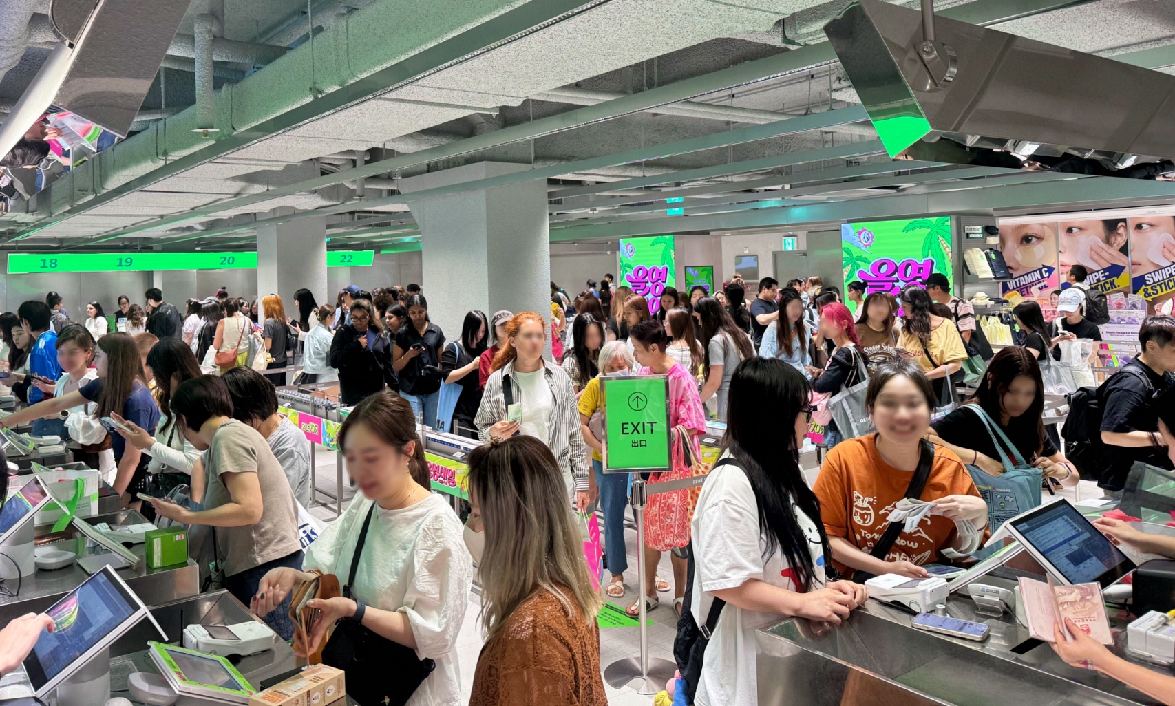 Crowds of shoppers queue at checkout counters beneath a green EXIT sign in a cosmetics store.