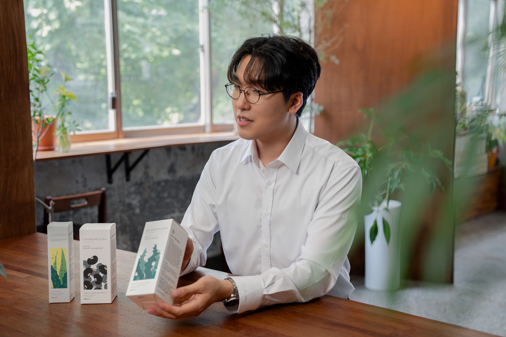 안경을 쓴 남성이 실내 나무 테이블에서 제품 상자를 들고 있으며 옆에 상자 두 개가 놓여 있다.