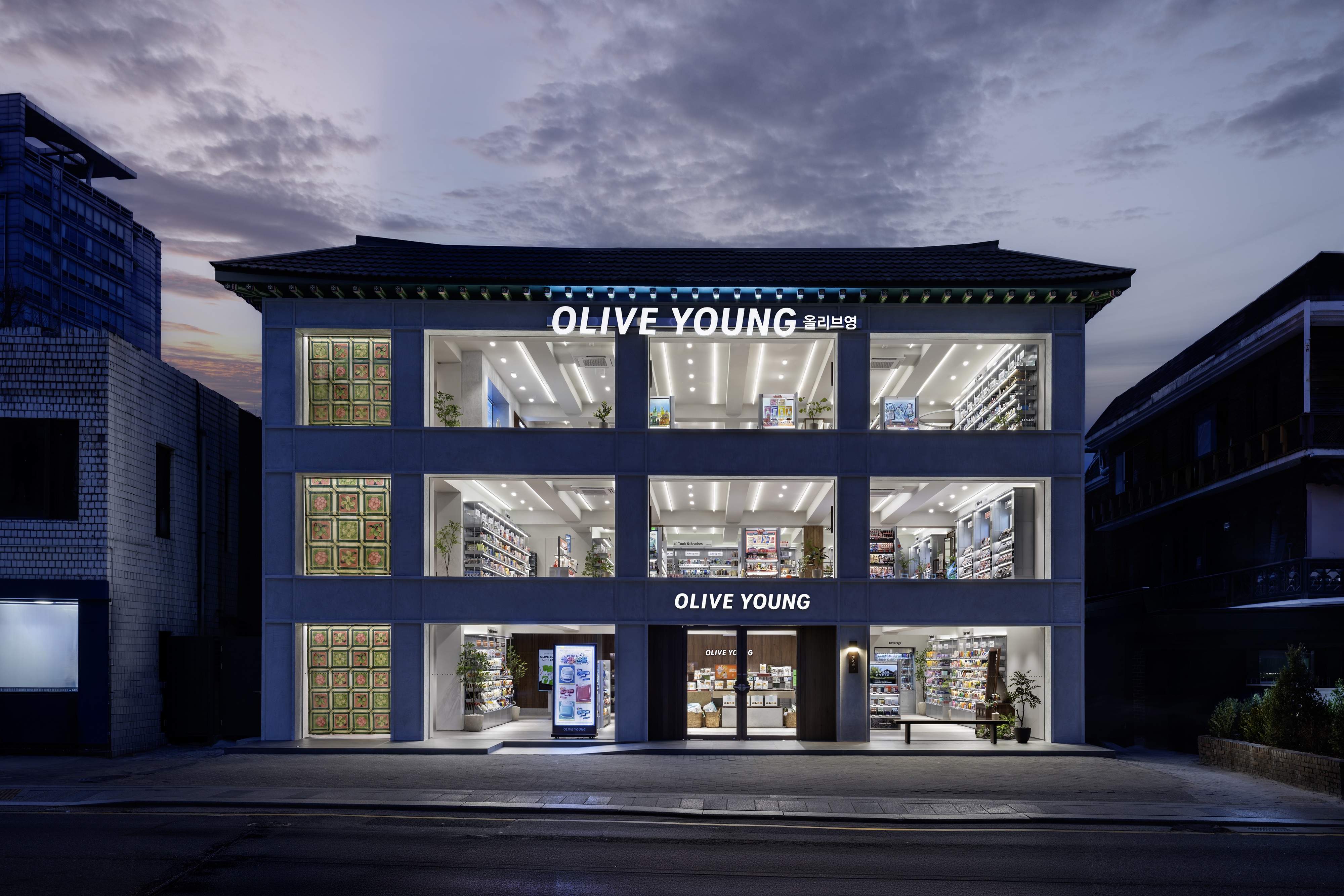 A brightly lit OLIVE YOUNG store occupies a traditional-roofed building at dusk.