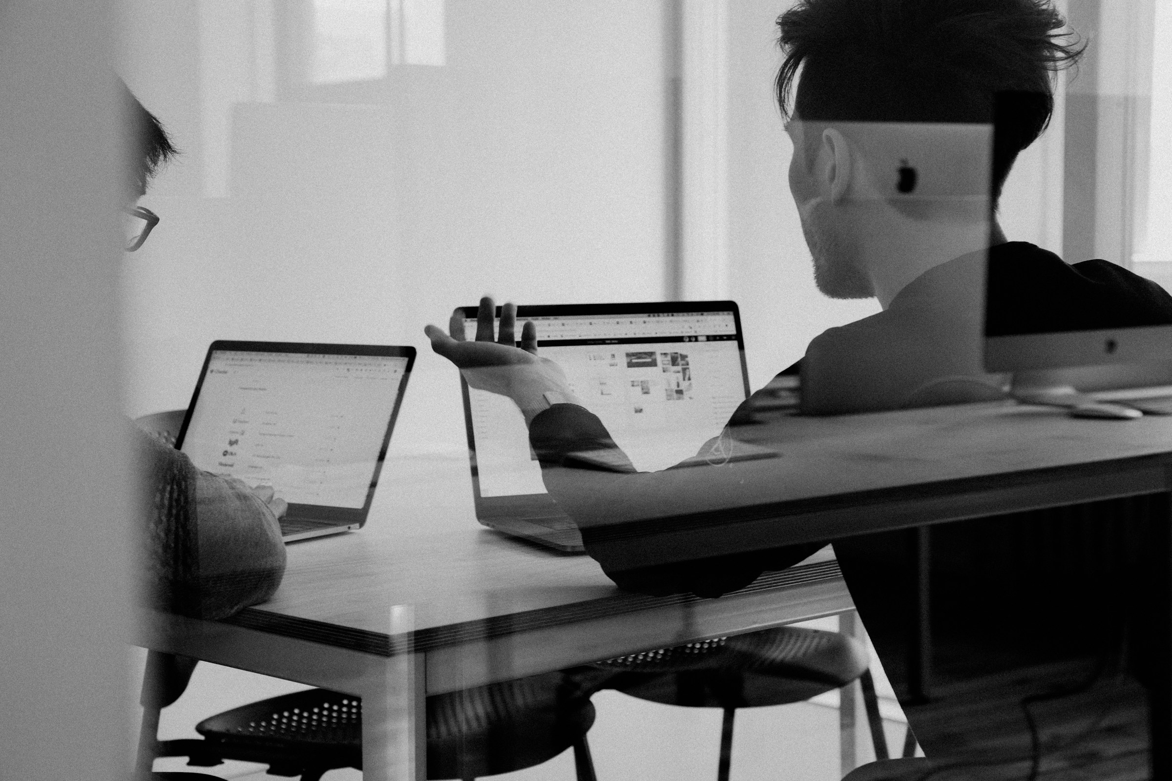2人の人物がテーブルを囲み、ノートパソコンを見ながら話し合っている
