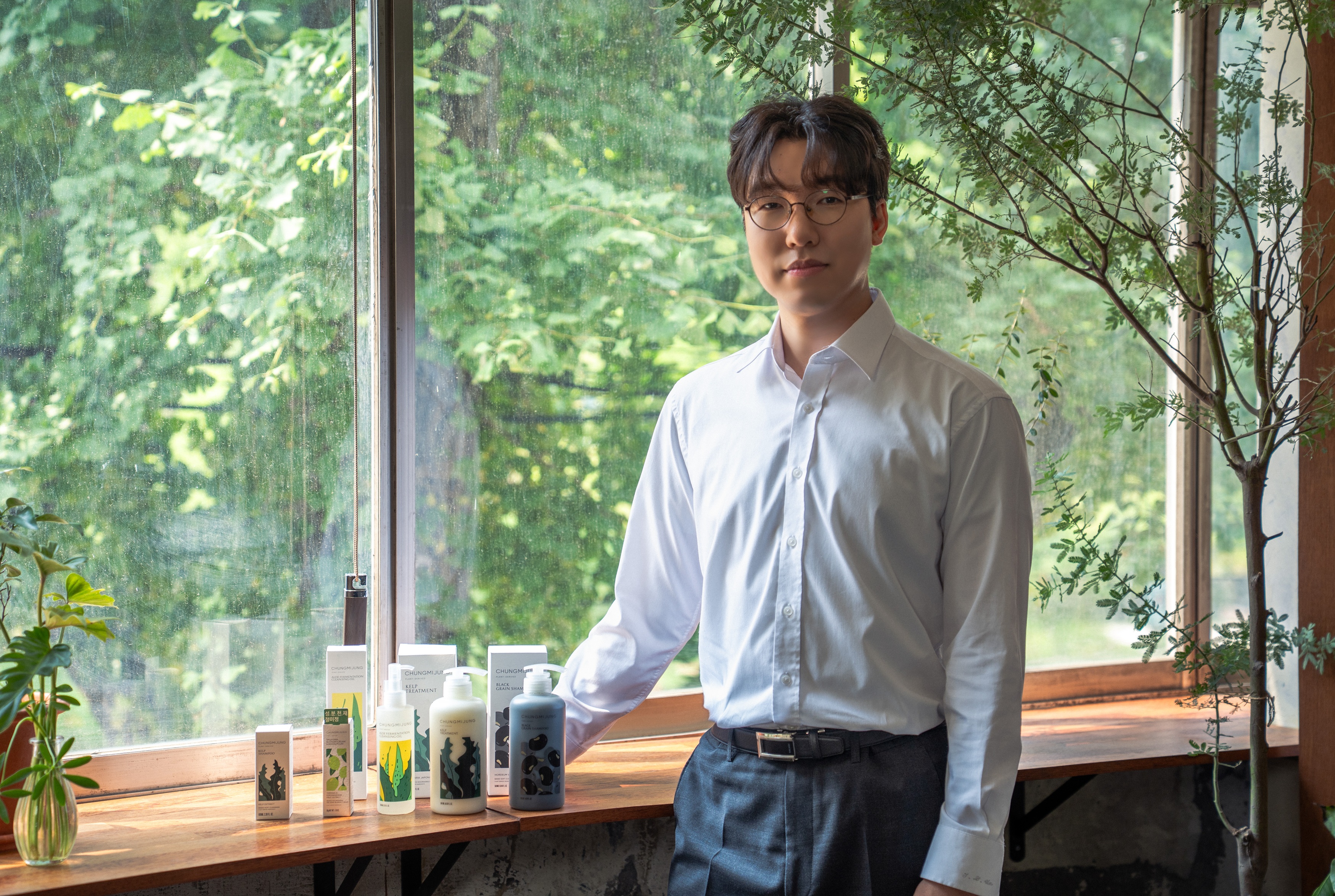 A man in glasses and a white shirt stands beside skincare and haircare products.