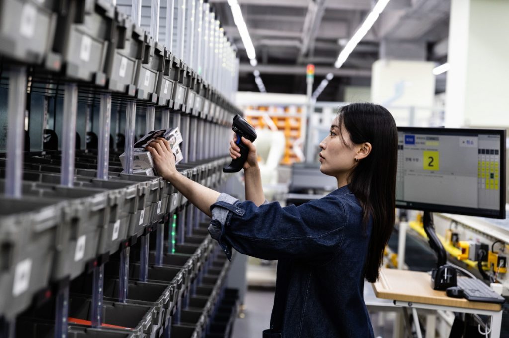 A woman scans a box with a handheld reader beside rows of storage bins.