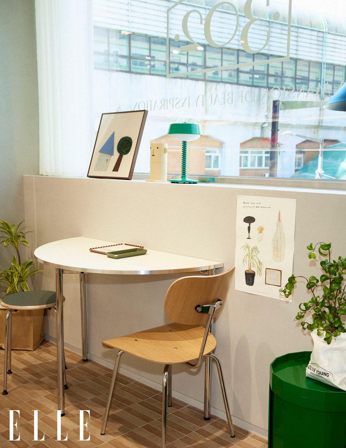 A bright corner has a round table, wooden chair, plants, artwork and a green lamp by a window.
