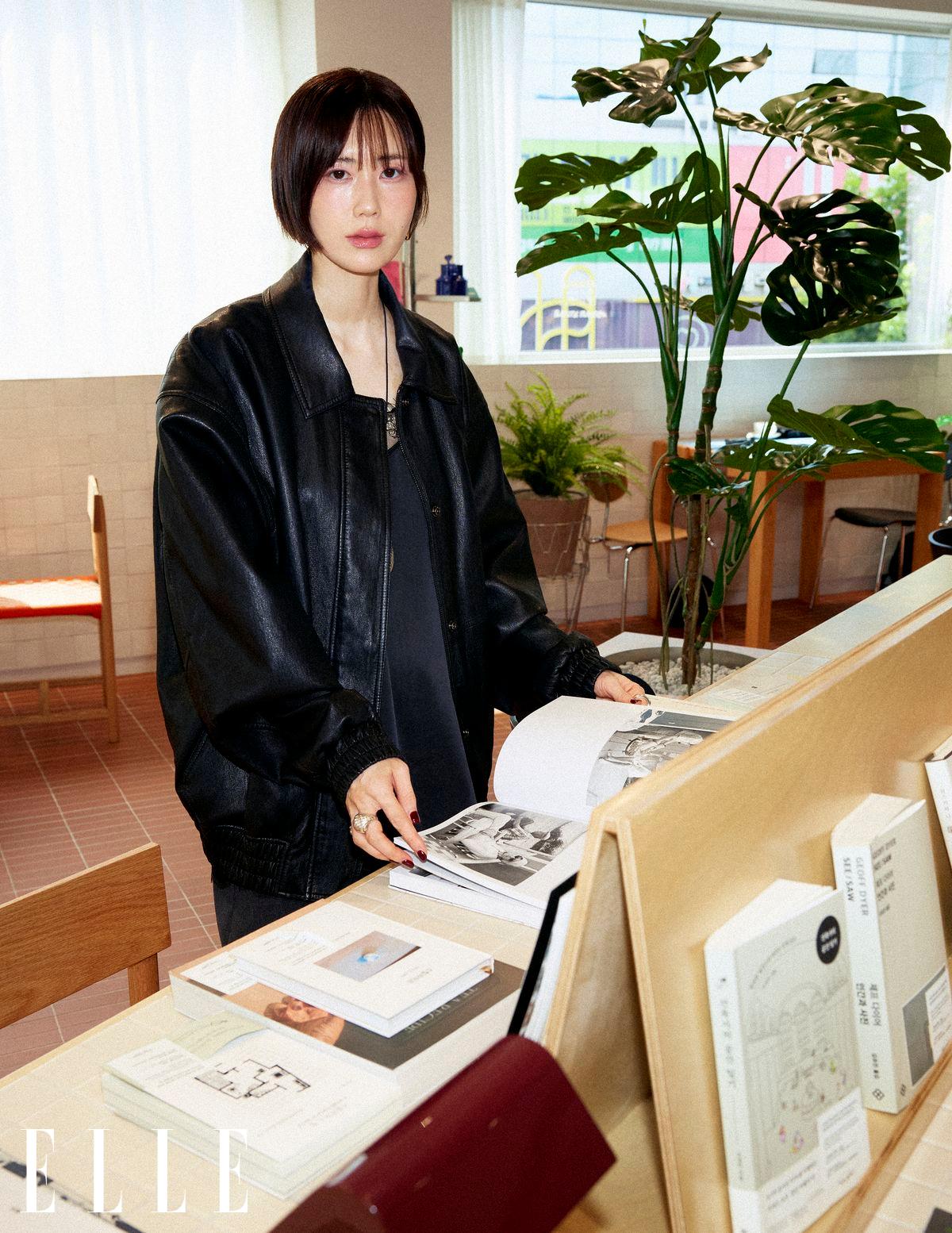 검은 가죽 재킷을 입은 여성이 책상에서 펼쳐진 책을 살펴보고 있다.
