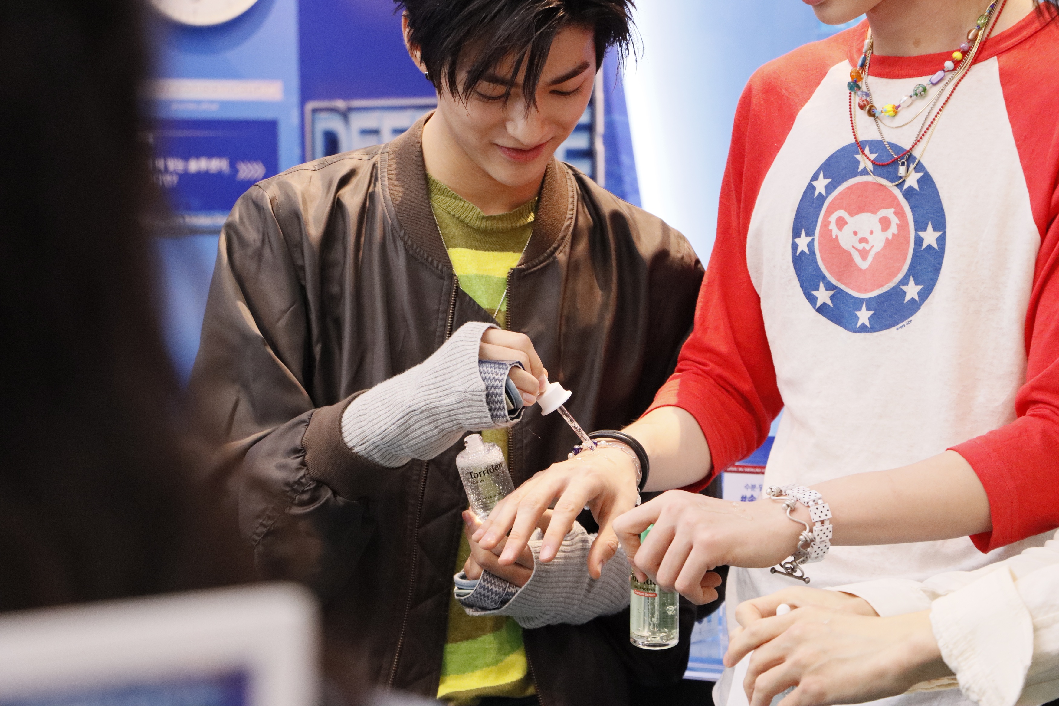 Two people handle skincare bottles as one uses a dropper on the other's hand.