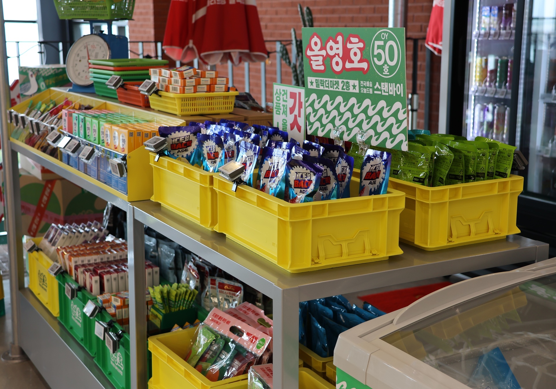 A shop display holds colorful packaged drinks and snacks in yellow crates beneath Korean signs.