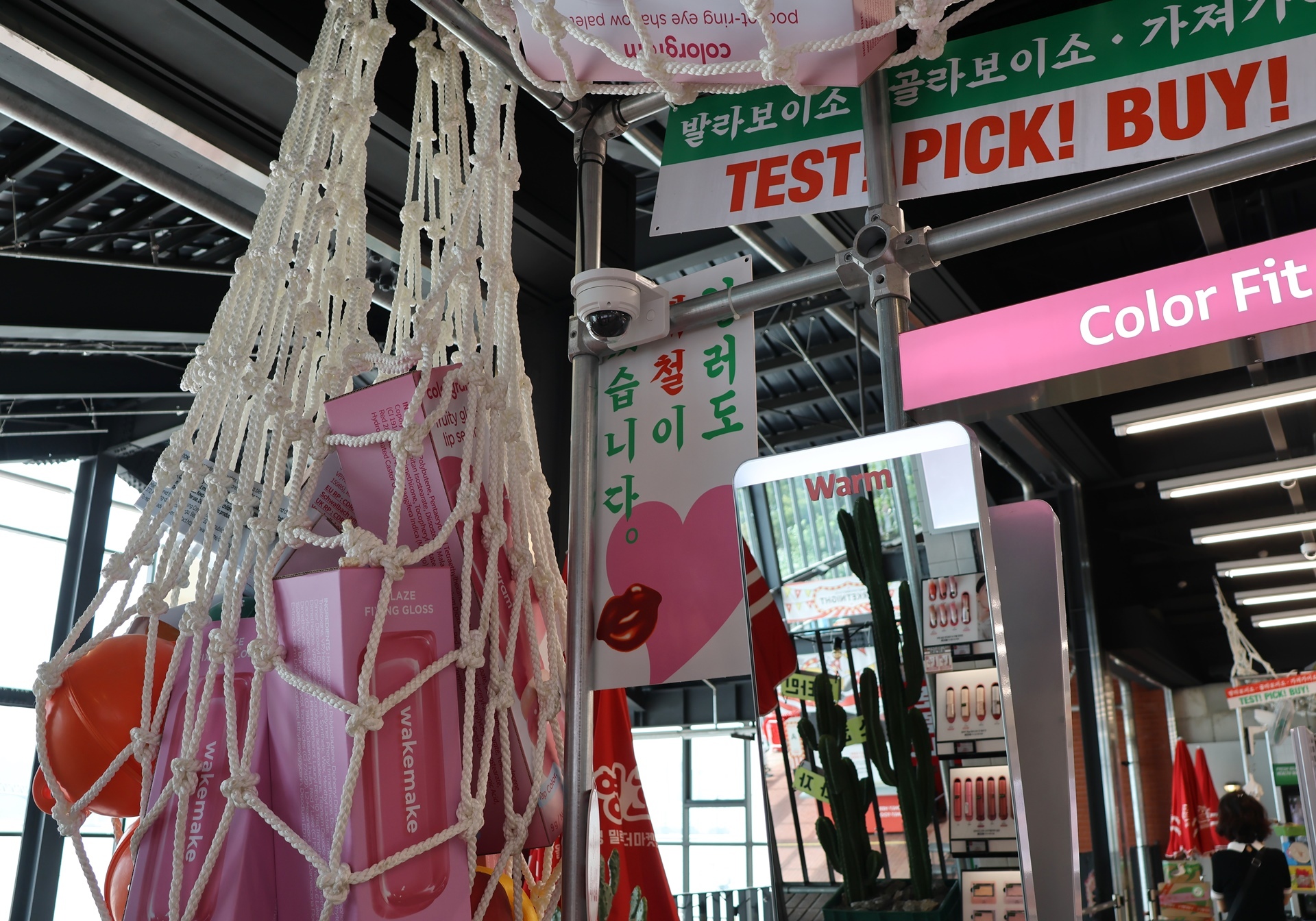 A net of pink cosmetics hangs beside a mirror and signs reading “TEST, PICK! BUY!”