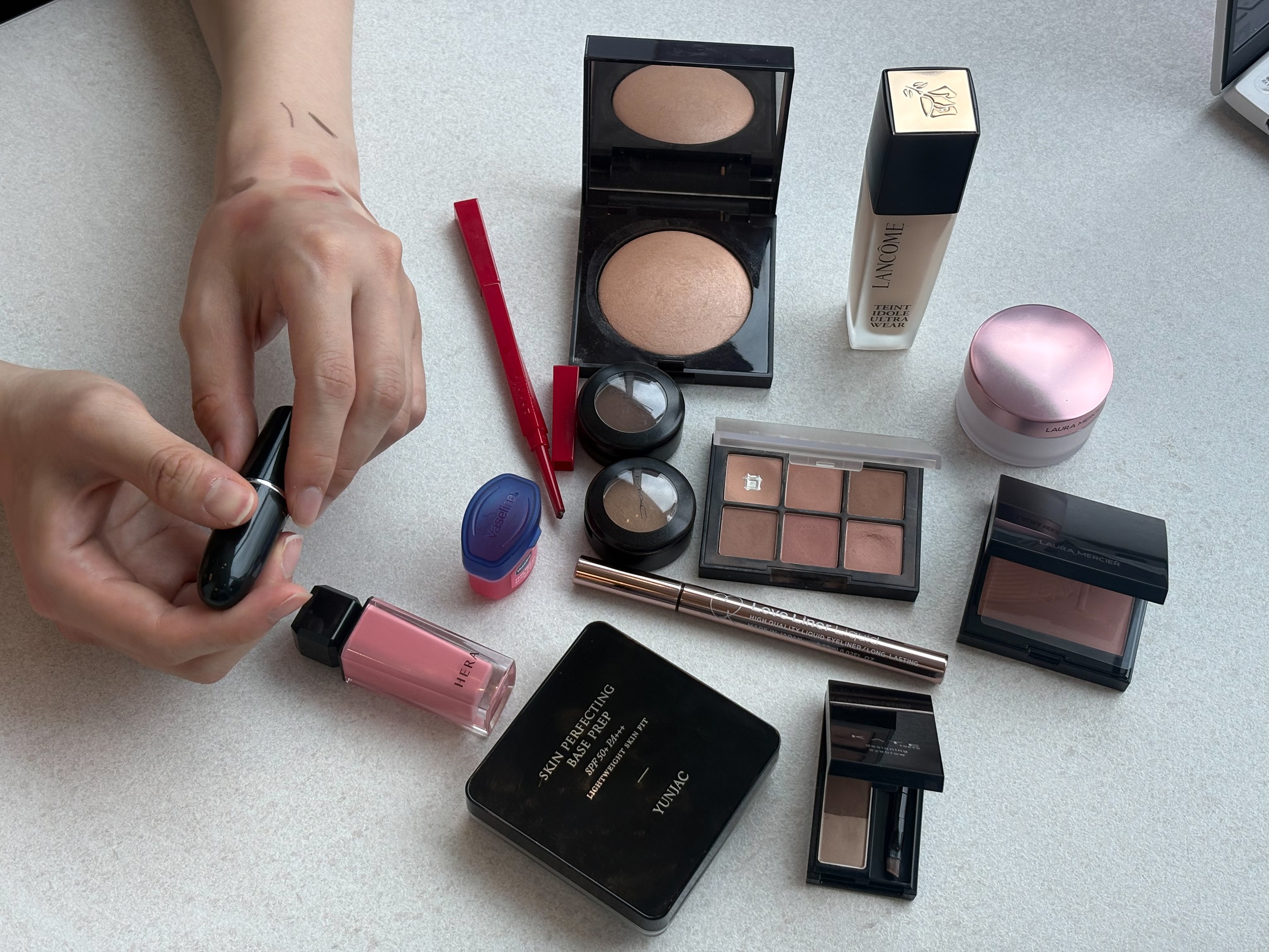 A hand holds a black makeup tube among assorted cosmetics on a tabletop.