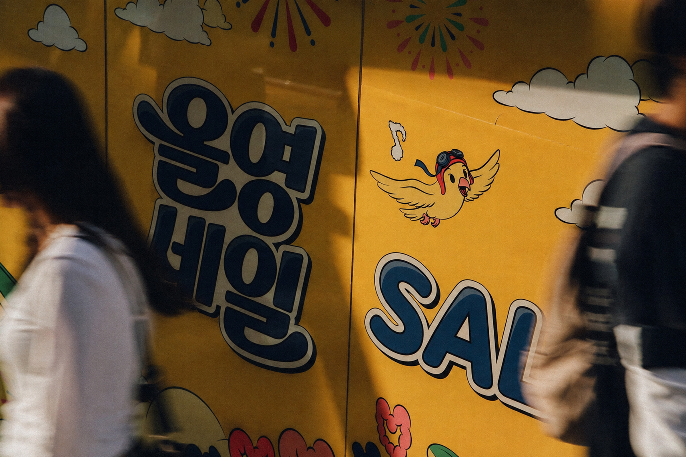 Blurred people pass a colorful mural featuring a flying cartoon bird and clouds.