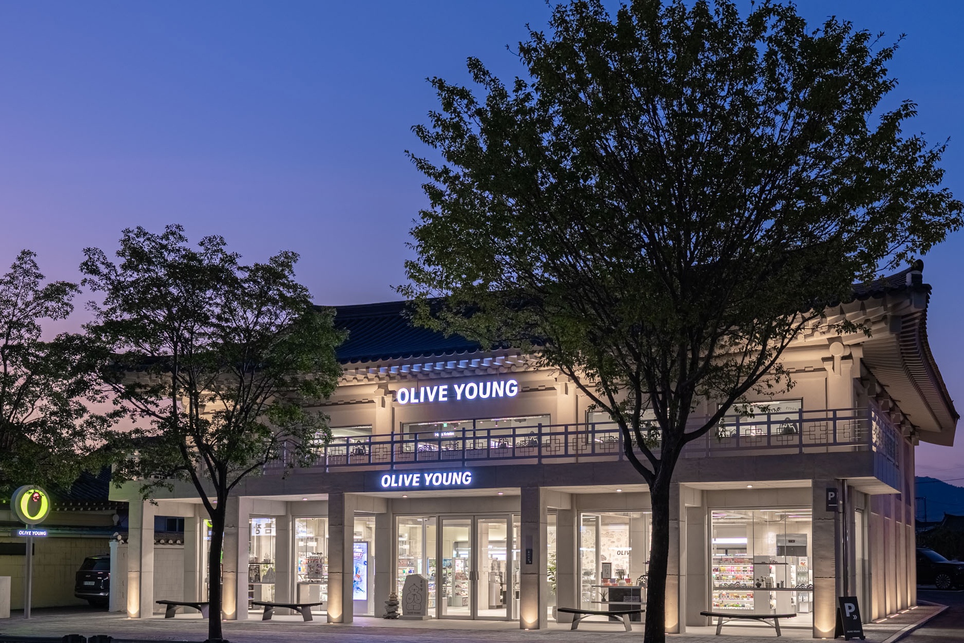 An Olive Young store with a traditional tiled roof is lit among trees at dusk.