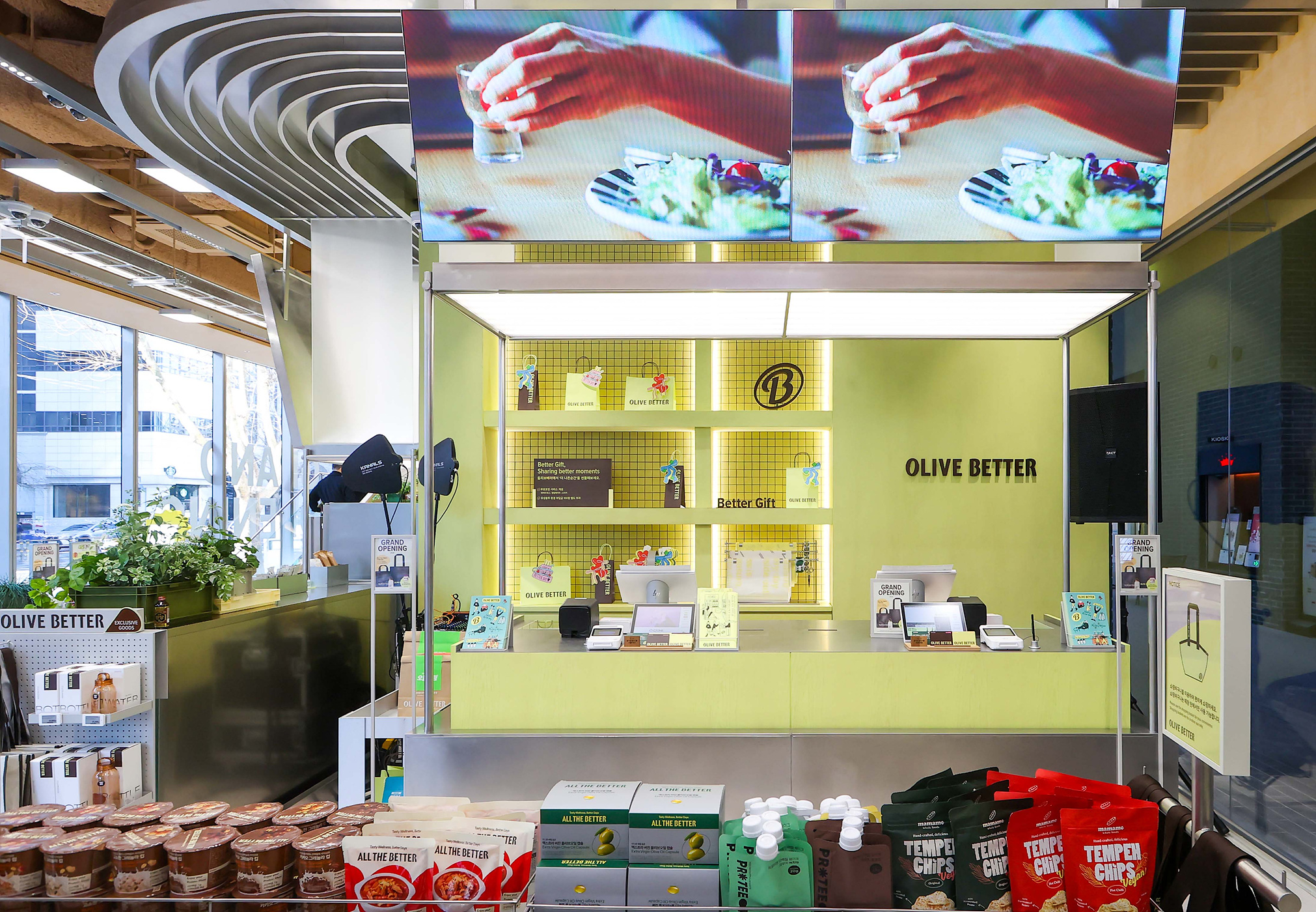 「OLIVE BETTER」の黄緑色の売り場に、商品棚とレジ、飲食映像を映す大型モニターがある。