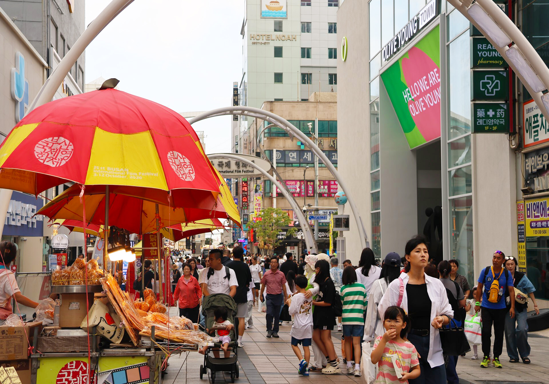 A crowded Korean shopping street features a red-and-yellow snack cart and arched decorations.