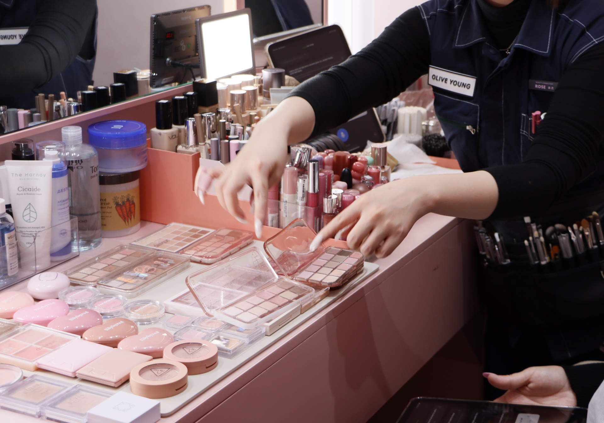 A makeup artist arranges eyeshadow palettes and cosmetics at a crowded vanity.