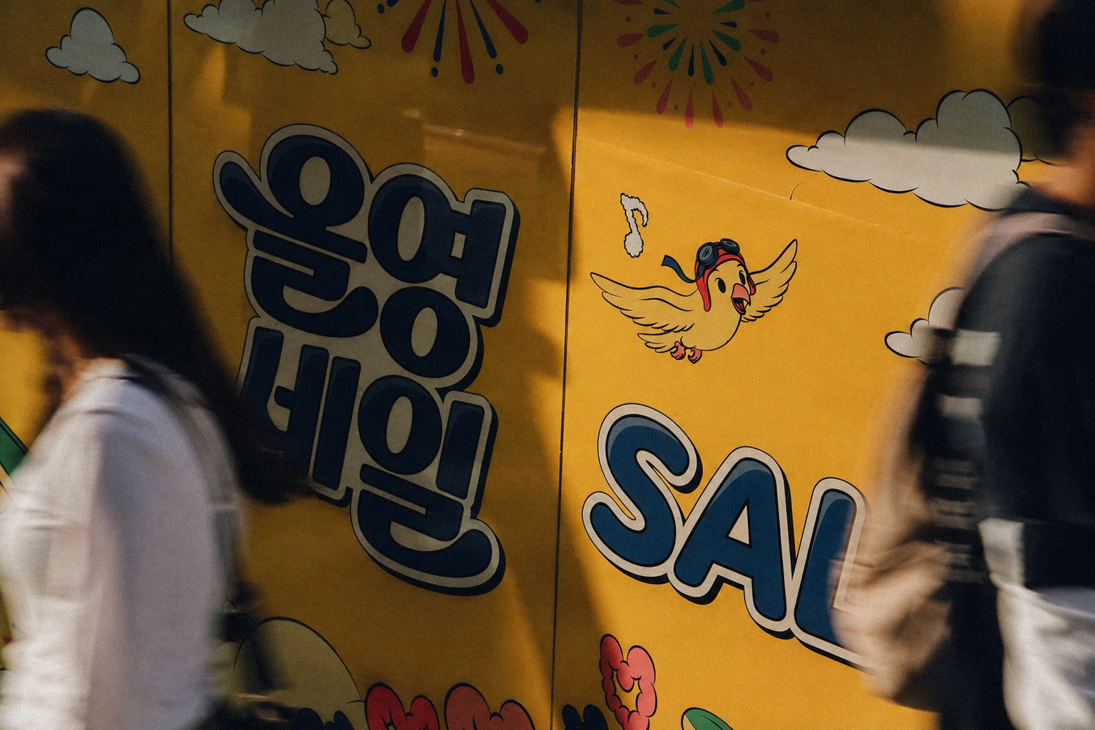 노란 벽에 구름과 불꽃, 날개 달린 새 그림이 있고 양옆으로 사람들이 지나간다.