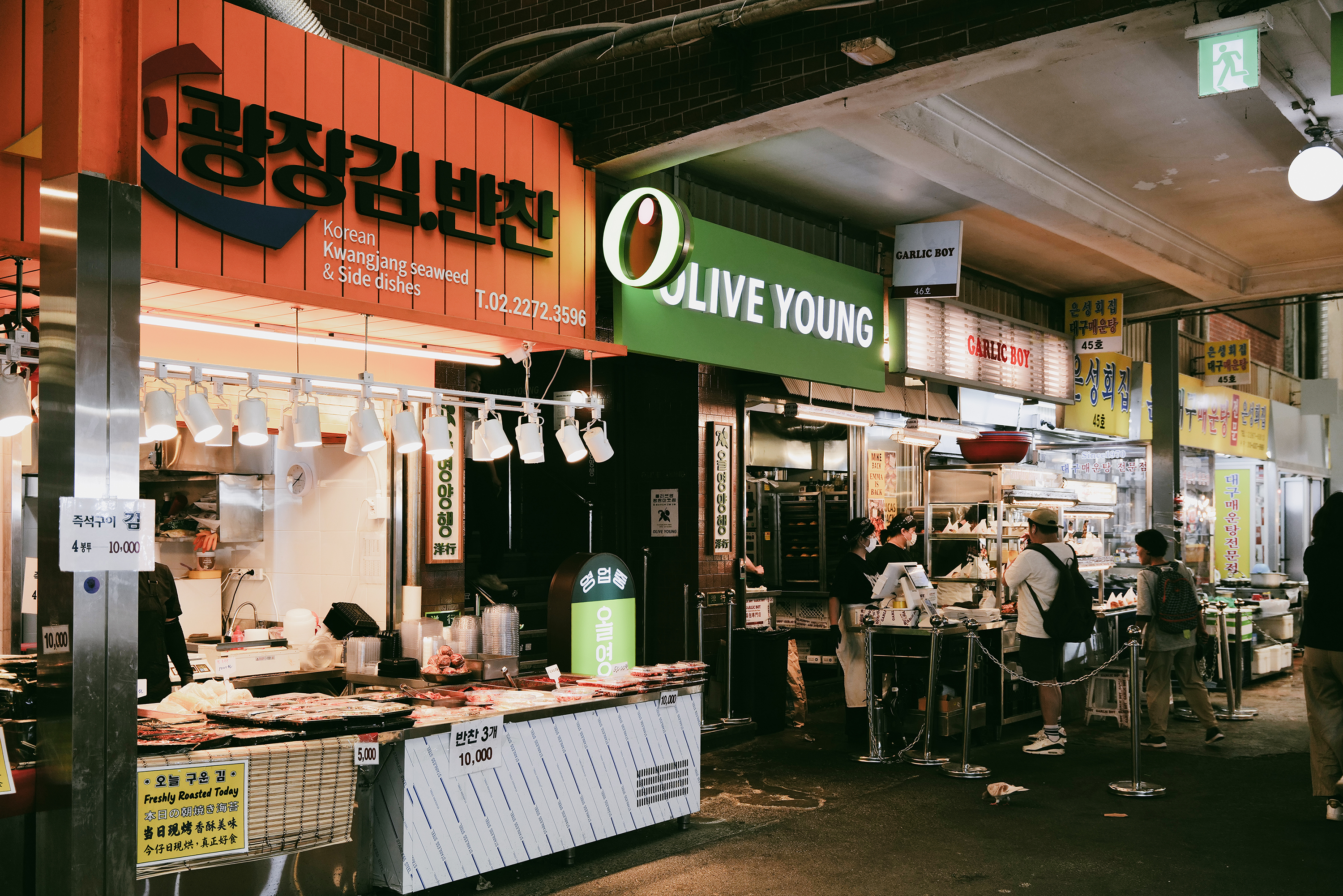 간판이 즐비한 시장 골목에서 상인들이 김과 음식을 판매하고 사람들이 오가고 있다.