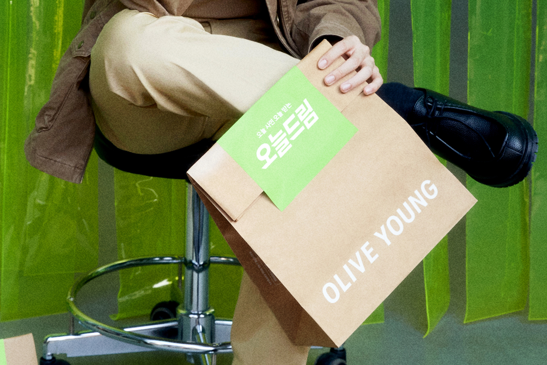 A seated person holds an OLIVE YOUNG same-day delivery bag over their lap.