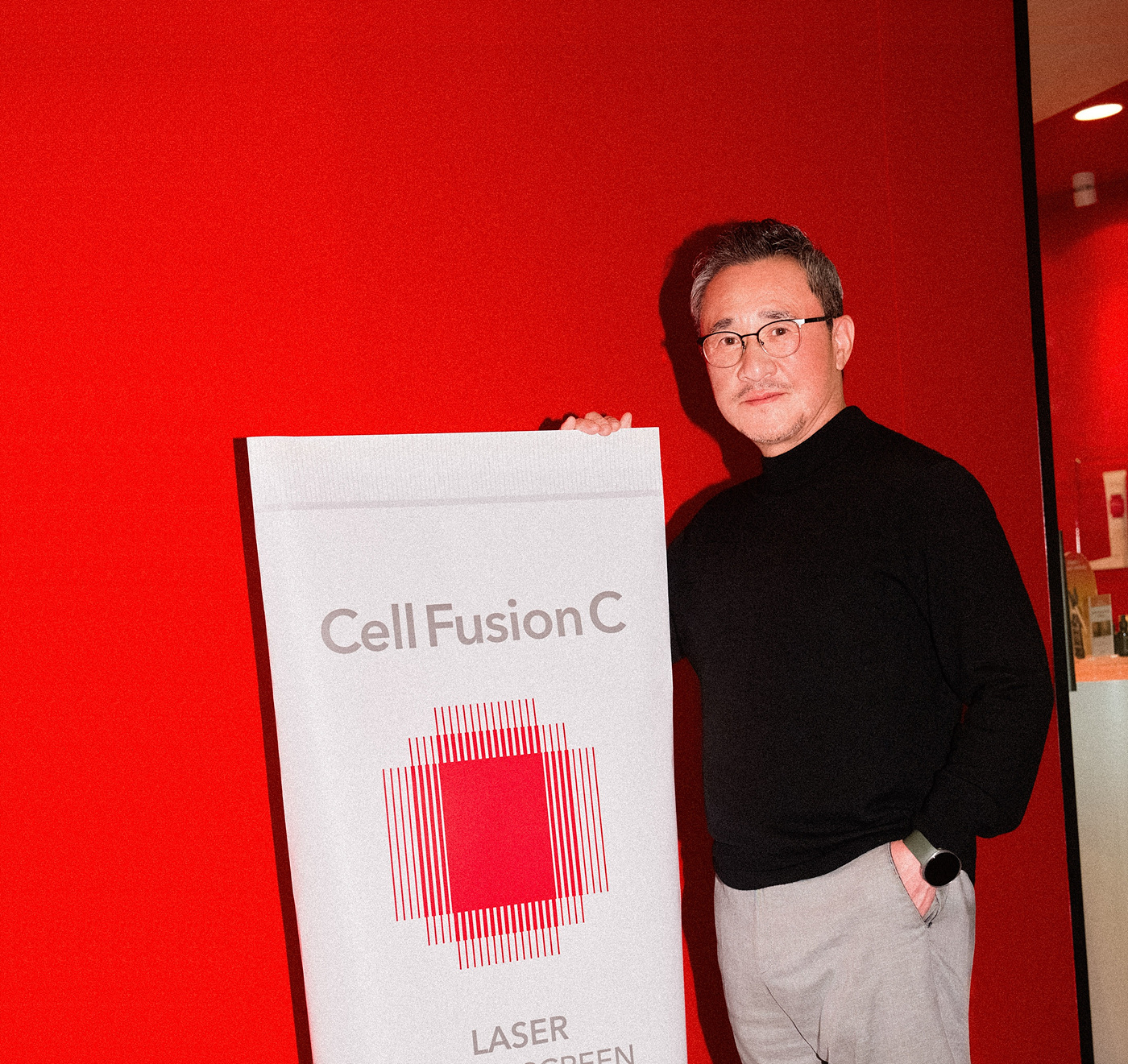 A man in glasses holds a “Cell Fusion C” poster beside a red wall.
